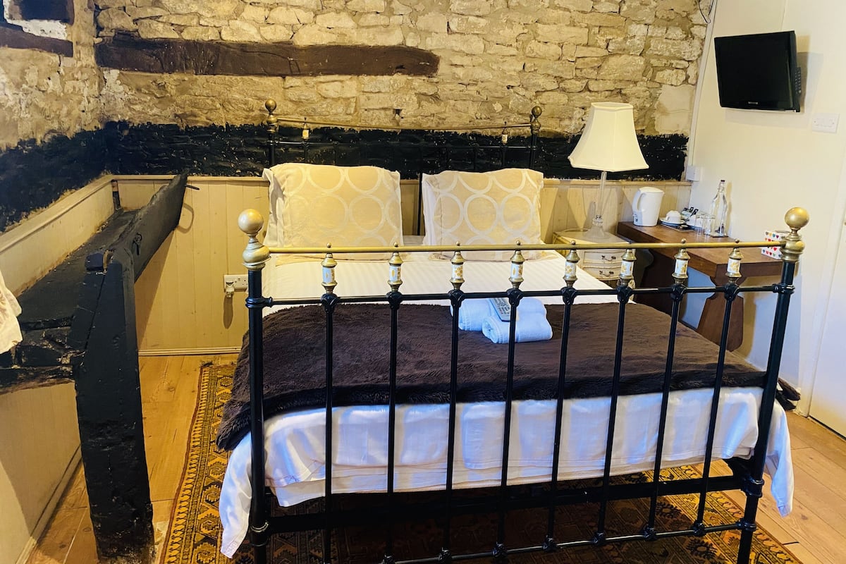 A room with a stone wall, a bed with a black metal frame, a wooden desk, and a television on the wall.