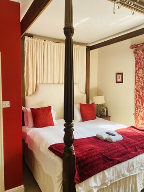 A four-poster bed with white linens and red pillows, a bedside table with a lamp and radio, and a red curtain. - The Old Court Hotel (Witney)