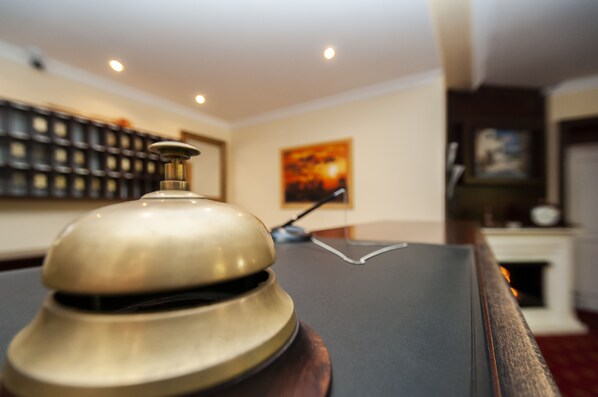 A hotel reception desk with a bell, a framed painting on the wall, and a bookshelf in the background.