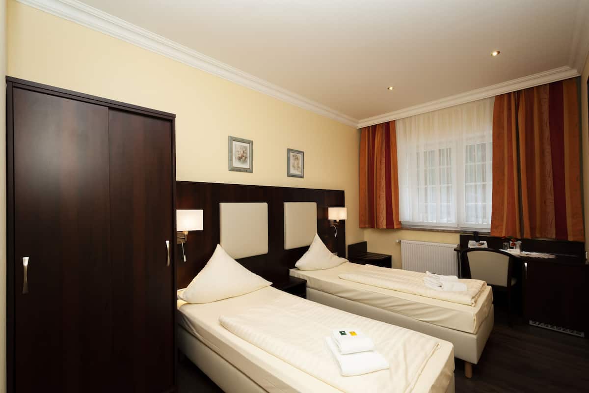 A hotel room with two single beds, a wooden wardrobe, a desk with a chair, and a window with curtains.