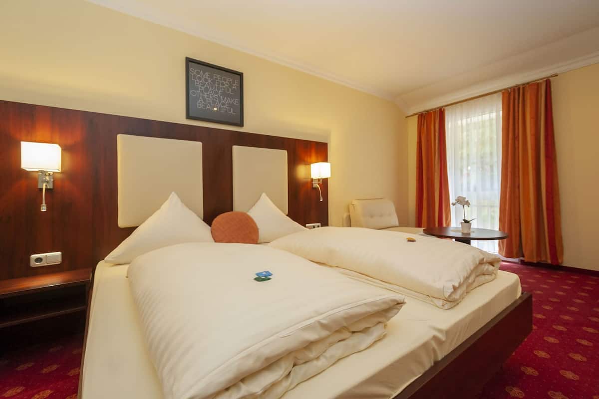 A hotel room with two beds, a wooden headboard, wall-mounted lamps, a small table with a plant, and a window with curtains.