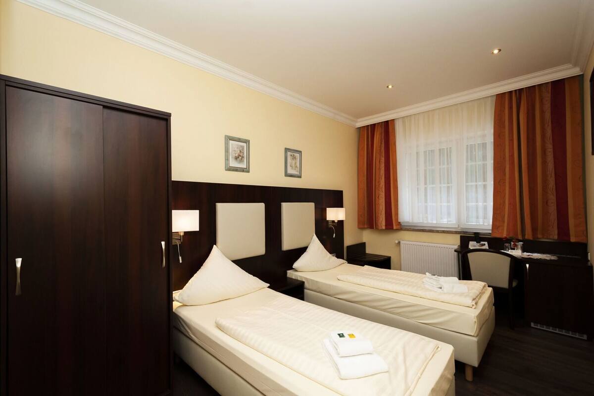 A hotel room with two beds, a desk, a wardrobe, and a window with curtains.