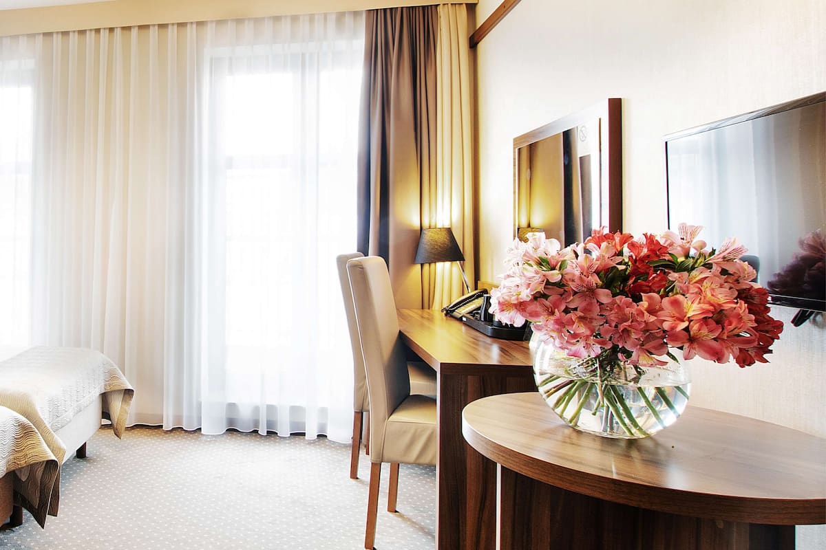 Una habitación de hotel con una cama, un escritorio con una lámpara, una silla y un florero con flores.