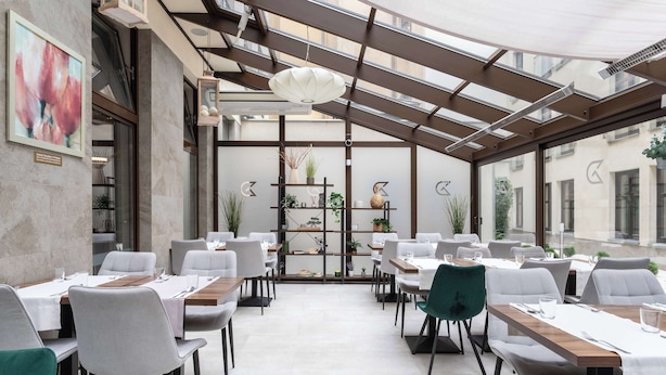 A modern restaurant with a glass ceiling, white tables, grey chairs, and a central plant display. - Golden Tulip Krakow City Center (Kraków)
