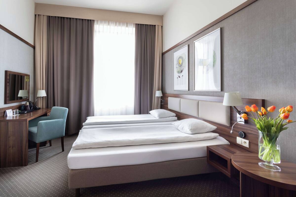 Habitación de hotel con cama, escritorio con silla, lámpara, florero con flores y una ventana grande con cortinas.