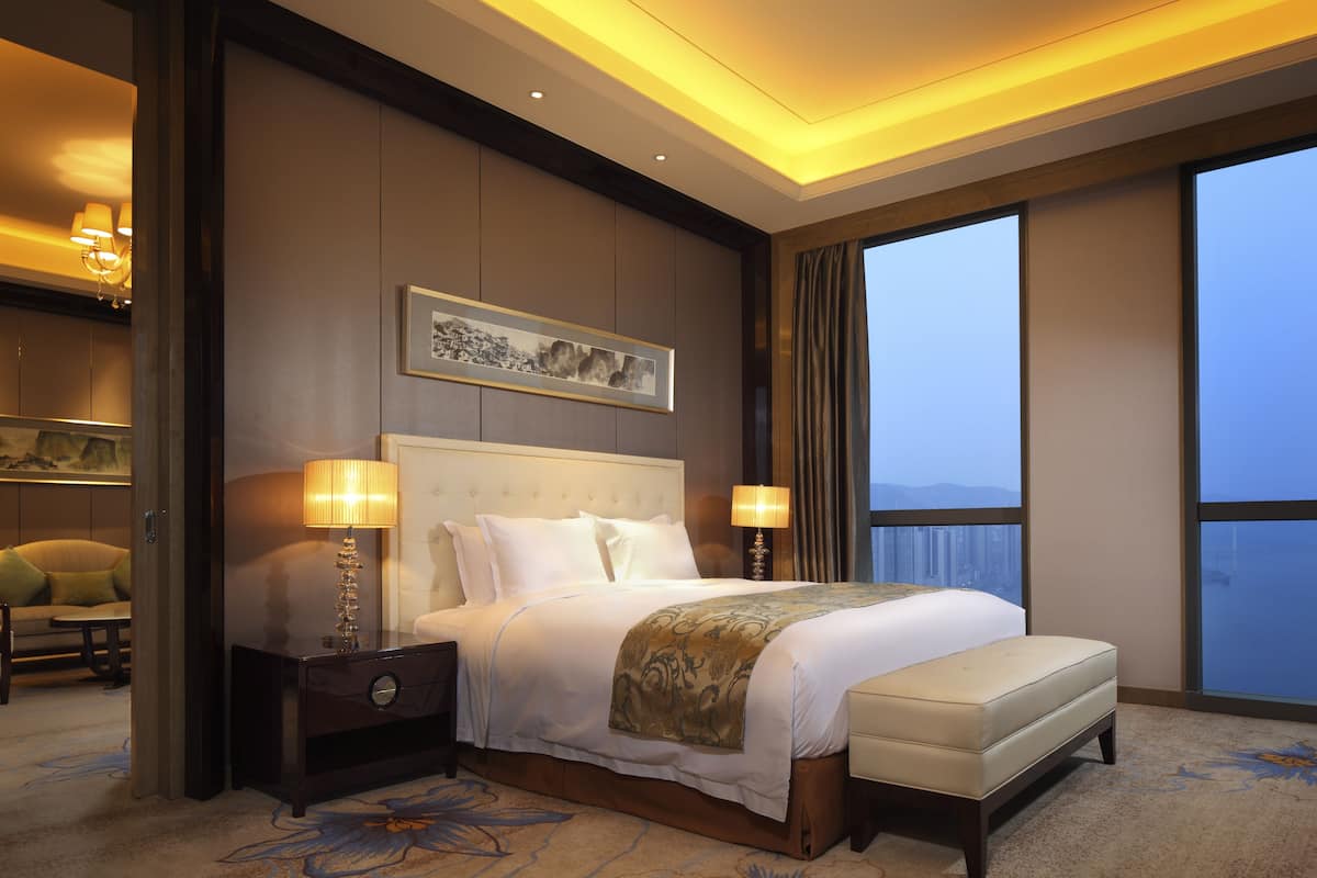 A hotel room with a large bed, two bedside tables with lamps, a headboard with a framed picture, and a view of the city through large windows.