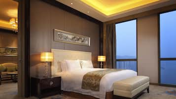 A hotel room with a large bed, two bedside tables with lamps, a headboard with a framed picture, and a view of the city through large windows.