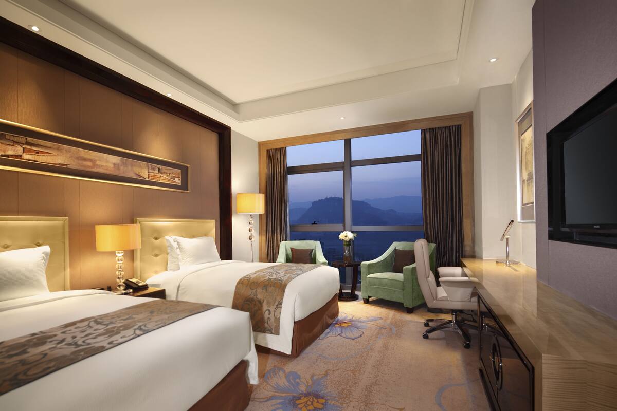 A hotel room with two beds, a desk, a TV, and a large window with a view.