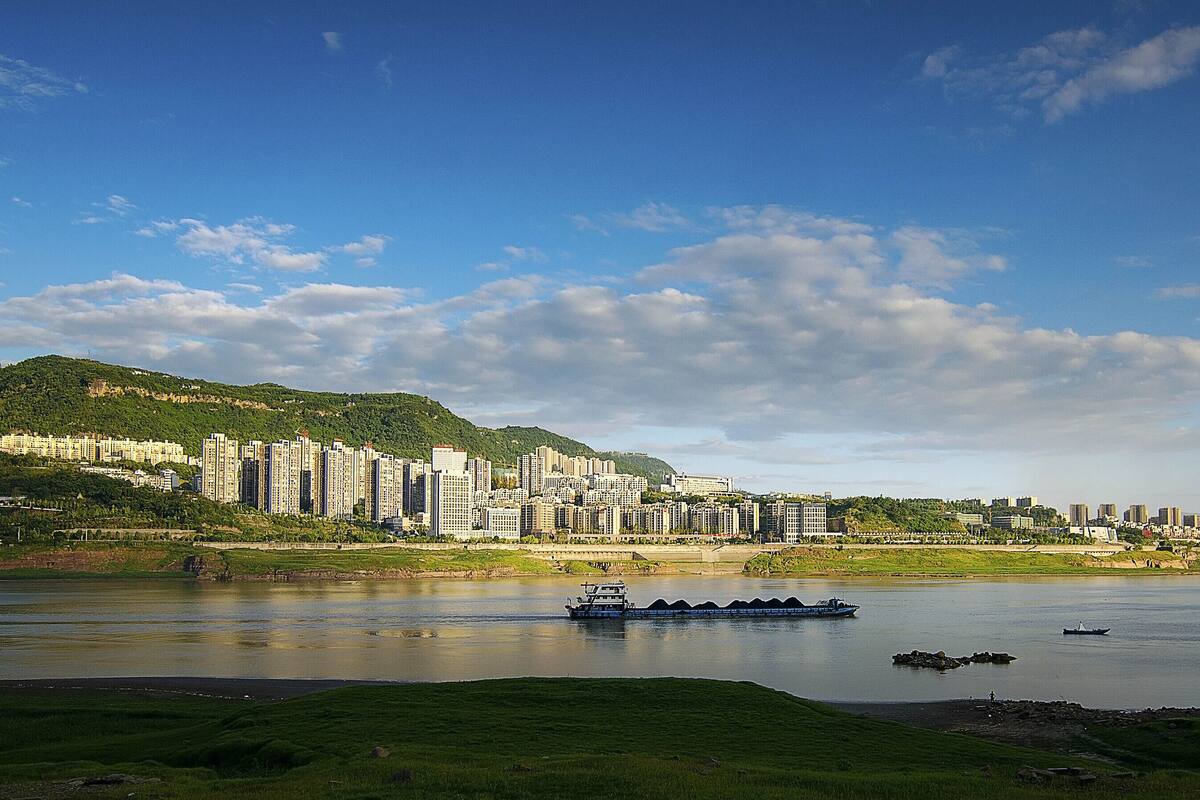 A river with boats, a cityscape with high-rise buildings, and a green hill.