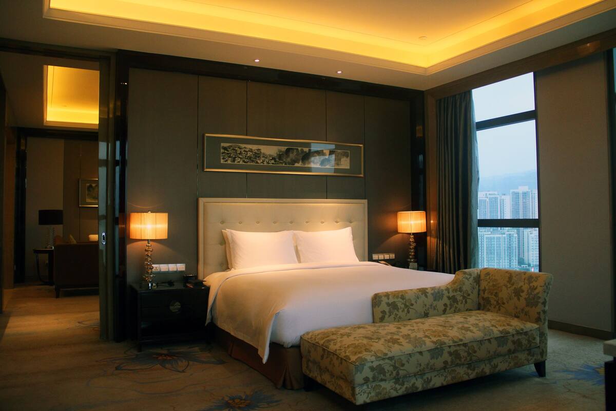 A hotel room with a large bed, two bedside lamps, a floral armchair, and a view of the city through a large window.