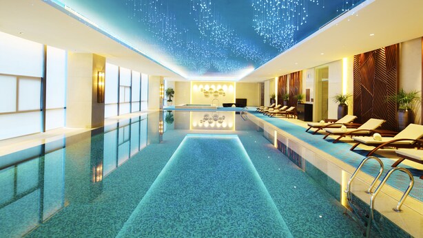 Un espace piscine intérieur moderne, doté d’un long bassin rectangulaire, de chaises longues et d’un plafond orné de luminaires suspendus.