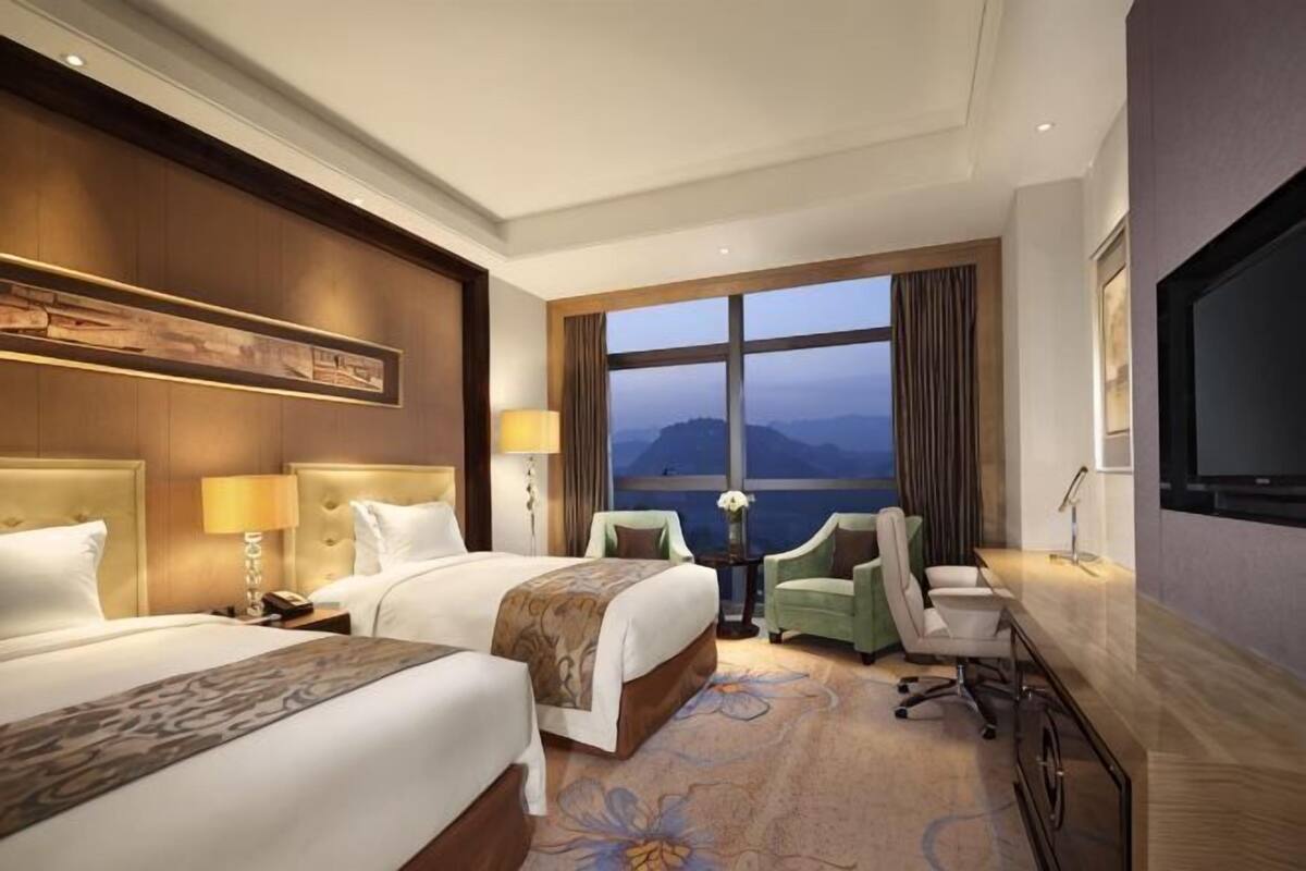 A hotel room with two beds, a desk, a chair, a TV, and a large window with a view.