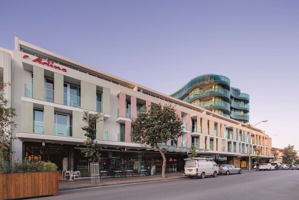 A modern hotel with a prominent 'Adina' sign, a covered outdoor seating area, and a curved glass building in the background. - Adina Apartment Hotel Bondi Beach Sydney (Bondi Beach)