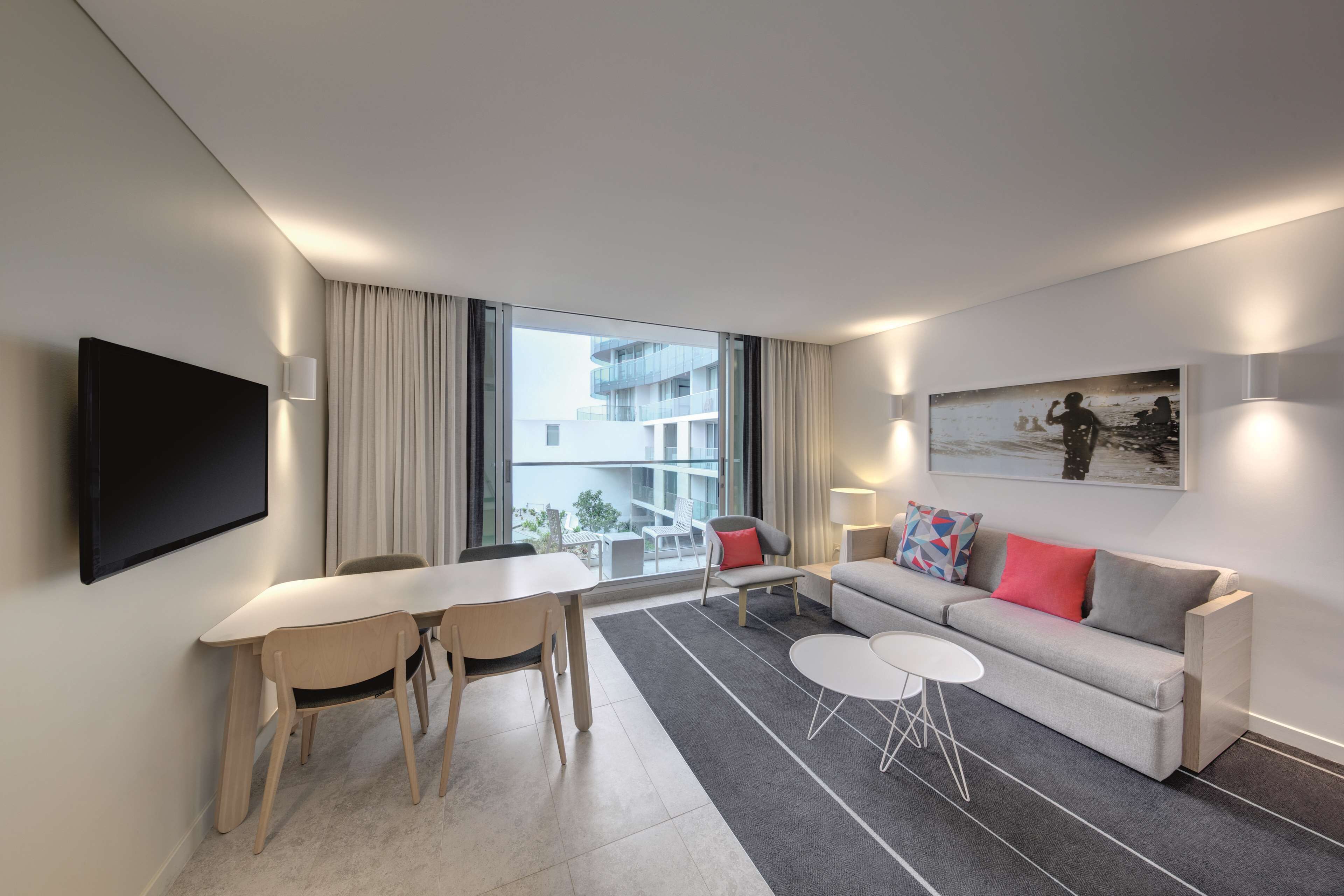 A modern hotel room with a dining area, a sofa, and a large window offering a view of the building.