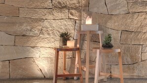 Dos taburetes de madera con velas, una piña y una planta pequeña frente a una pared de piedra.