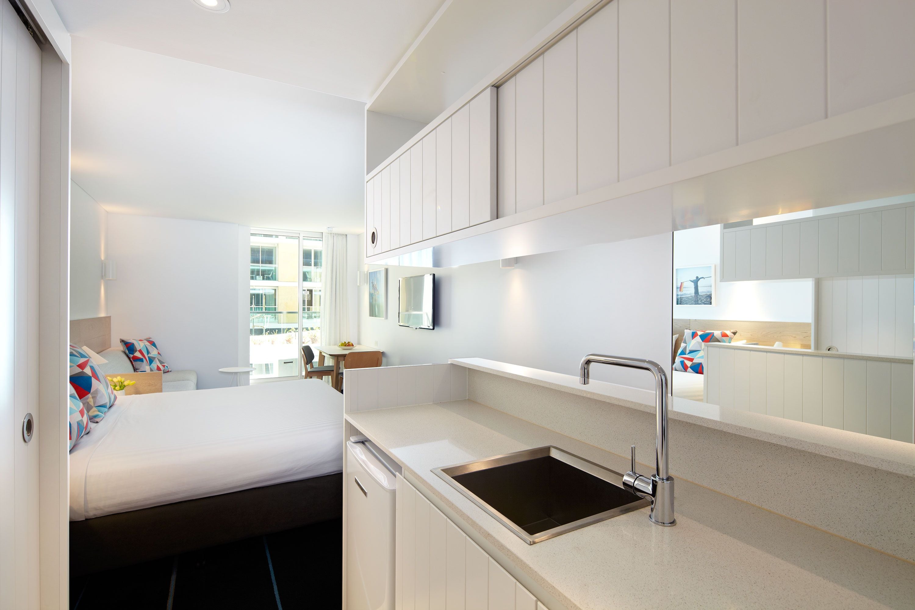 A compact hotel room with a bed, a small kitchenette, a sink, and a mirror.