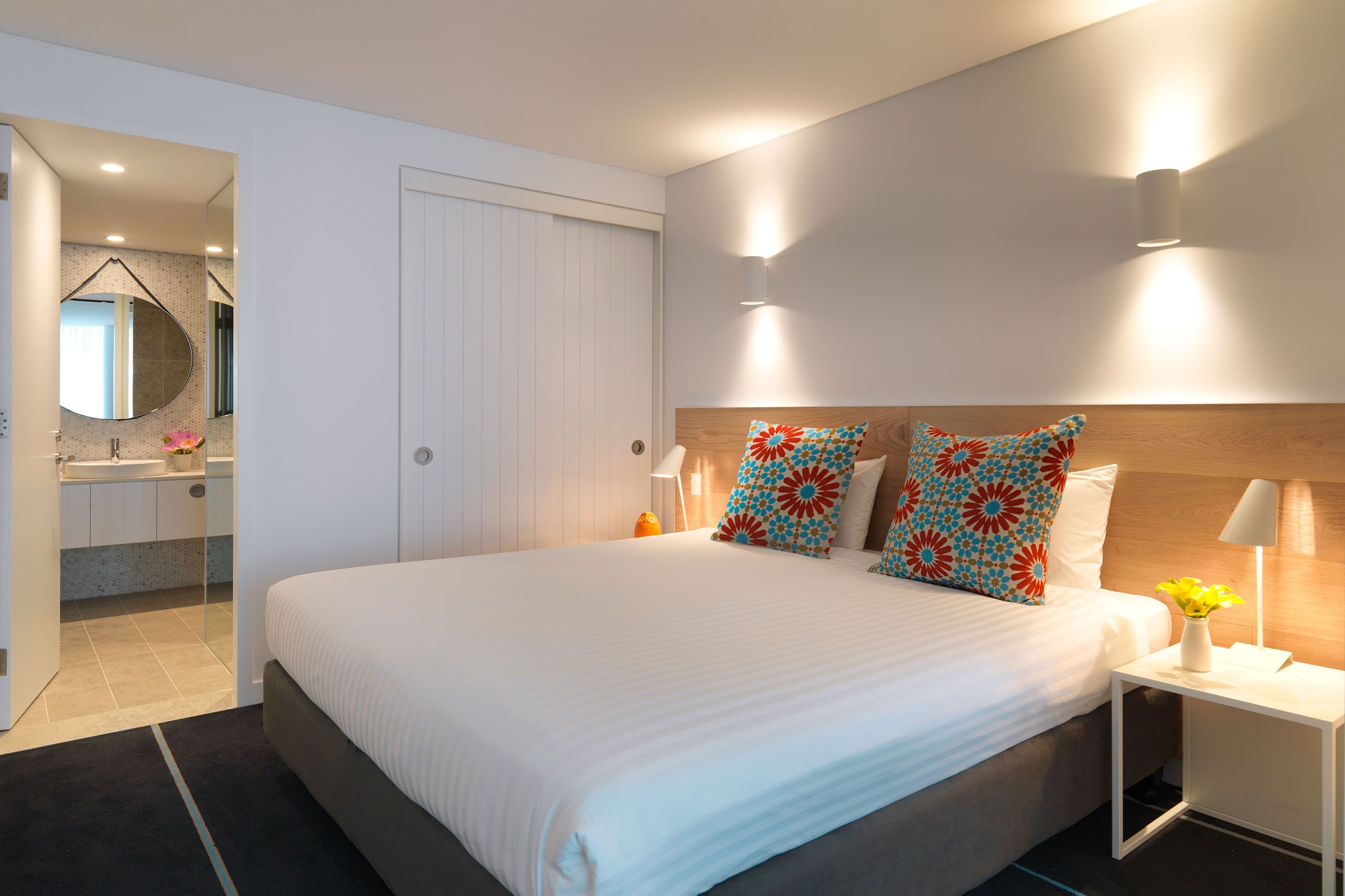 A modern hotel room with a large bed, a bedside table with a lamp, and a bathroom visible through an open door.