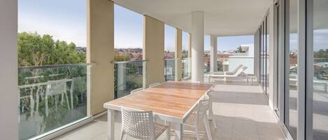Ein Balkon mit Holztisch und weißen Stühlen, großen Fenstern und Blick auf Gebäude und Bäume.