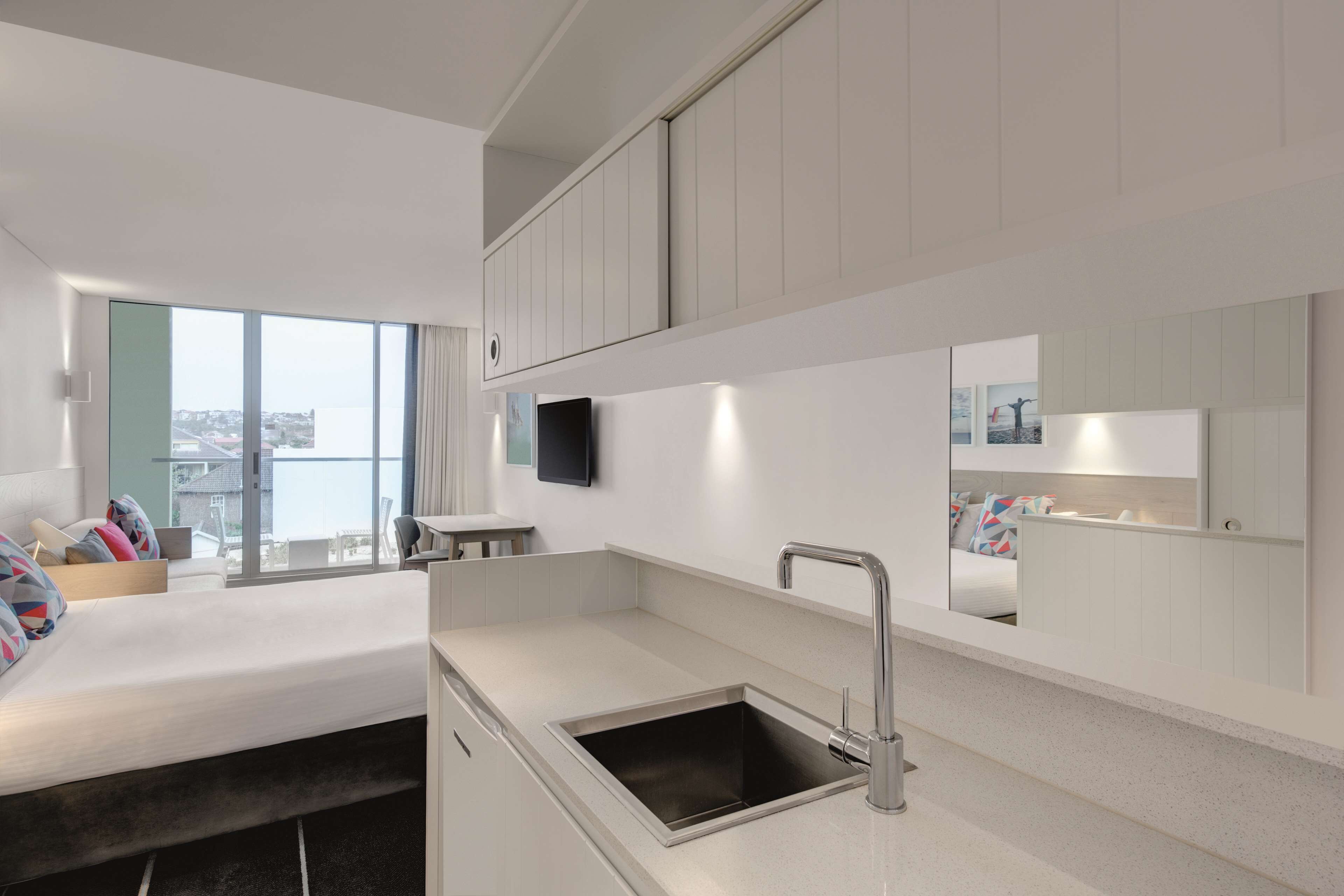 A modern hotel room with a large bed, a built-in desk, a small kitchen area with a sink, and a large window offering a city view.