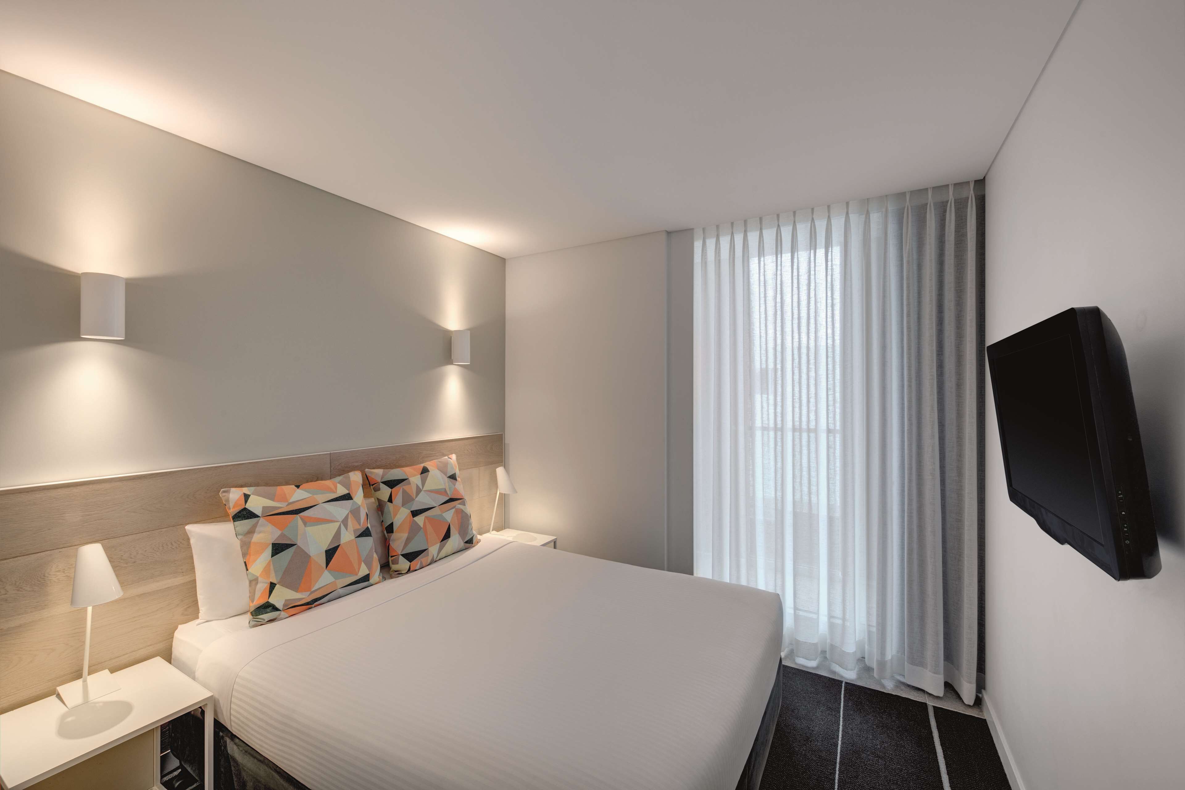 Ein modernes Hotelzimmer mit einem ordentlich bezogenen Bett, einem an der Wand befestigten Flachbildfernseher und einem Fenster mit durchscheinenden Vorhängen.