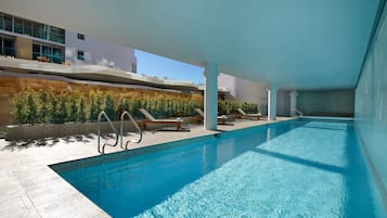 Una piscina interior moderna con agua azul cristalina, sillas de descanso y una pared de piedra con vegetación.