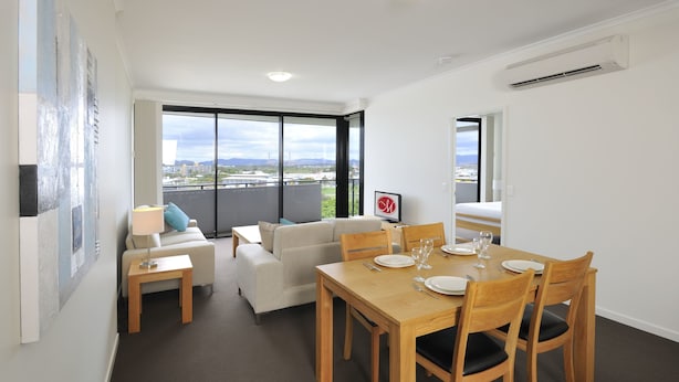 Un comedor moderno con una mesa de madera dispuesta para seis comensales, un sofá y vistas de la ciudad a través de amplios ventanales.