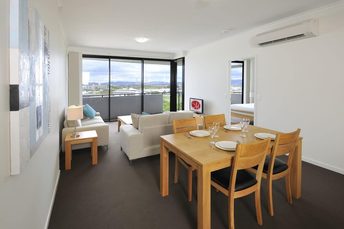 A modern dining area with a wooden table set for six, a sofa, and a view of the cityscape through large windows.