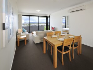 A modern dining area with a wooden table set for six, a sofa, and a view of the cityscape through large windows.
