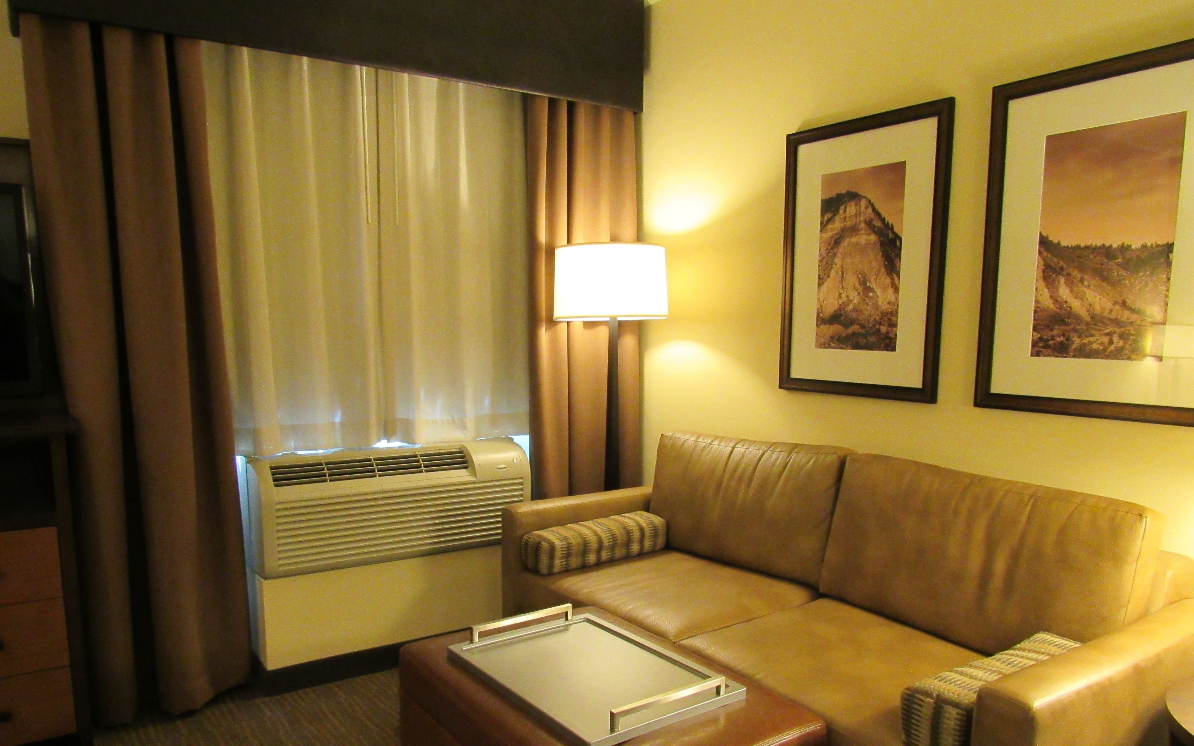 A hotel room with a leather sofa, a standing lamp, and two framed pictures on the wall.