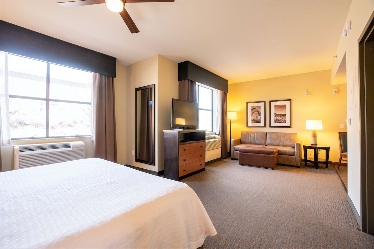 A hotel room with a large bed, a desk with a flat-screen TV, a sofa, a small table with a lamp, and a window with curtains.