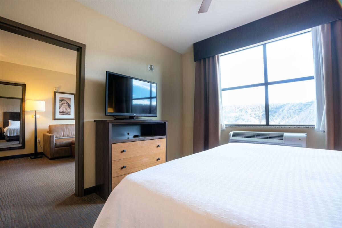 A hotel room with a large bed, a flat-screen TV on a wooden dresser, a window with curtains, and a view of snow-covered hills.