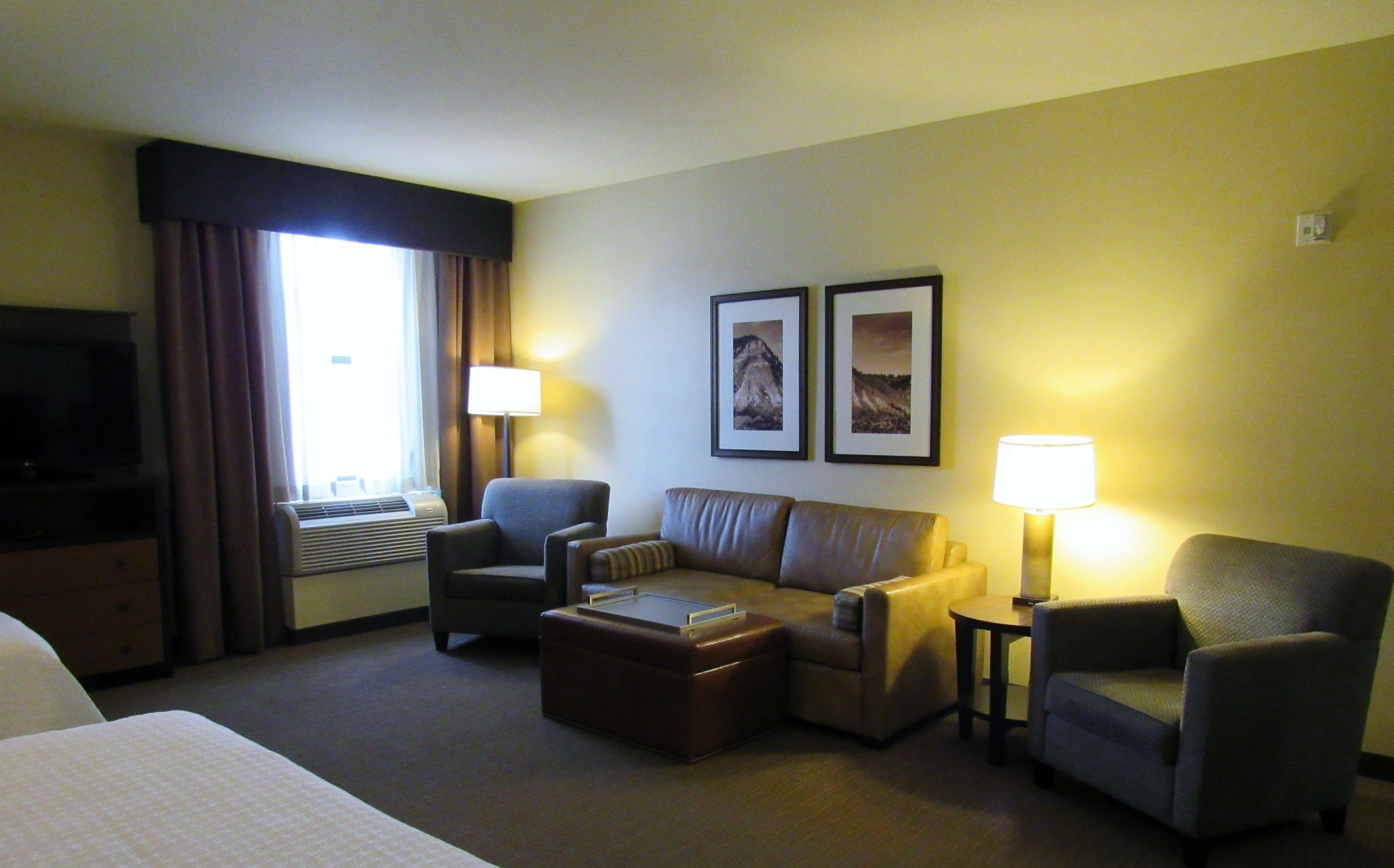 A hotel room with a bed, a sofa, two armchairs, a small table, a lamp, a TV, and two framed pictures on the wall.