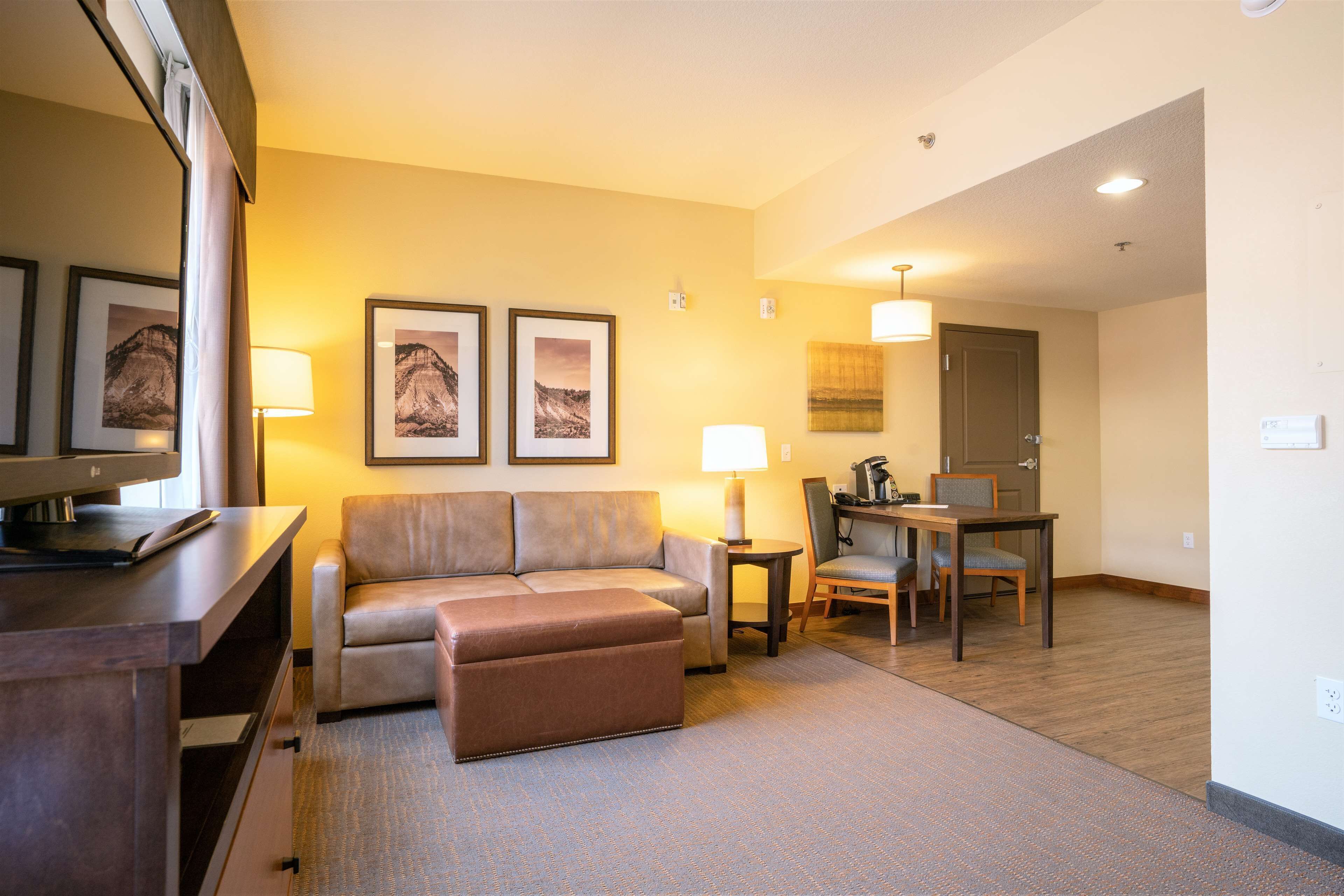 A hotel room with a sofa, ottoman, dining table, chairs, and artwork on the walls.