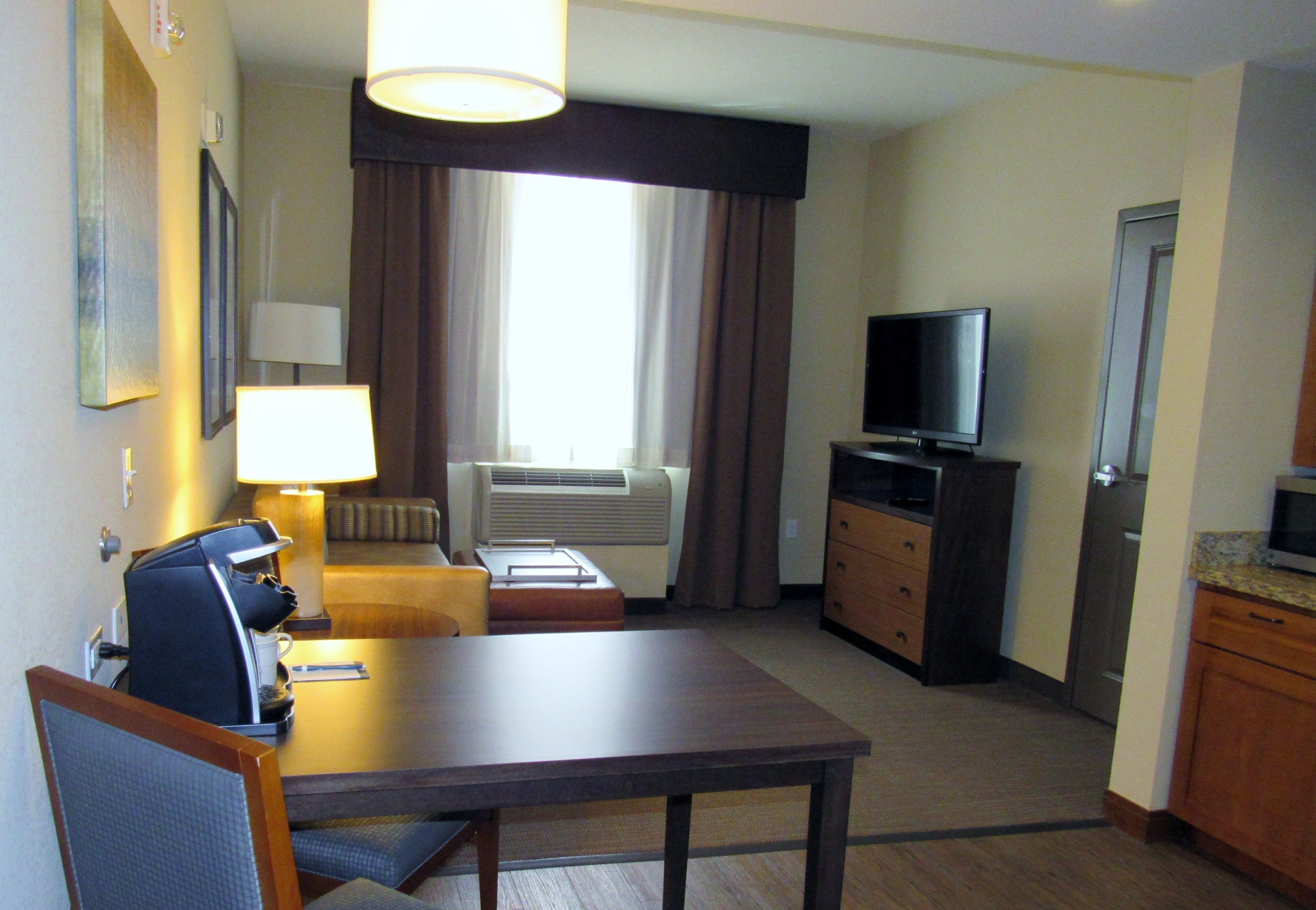 Habitación de hotel con una mesa de comedor, televisor, cafetera, sofá y ventana con cortinas.
