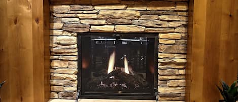 A stone fireplace with a wooden mantel, a fire burning inside, and a potted plant to the right.