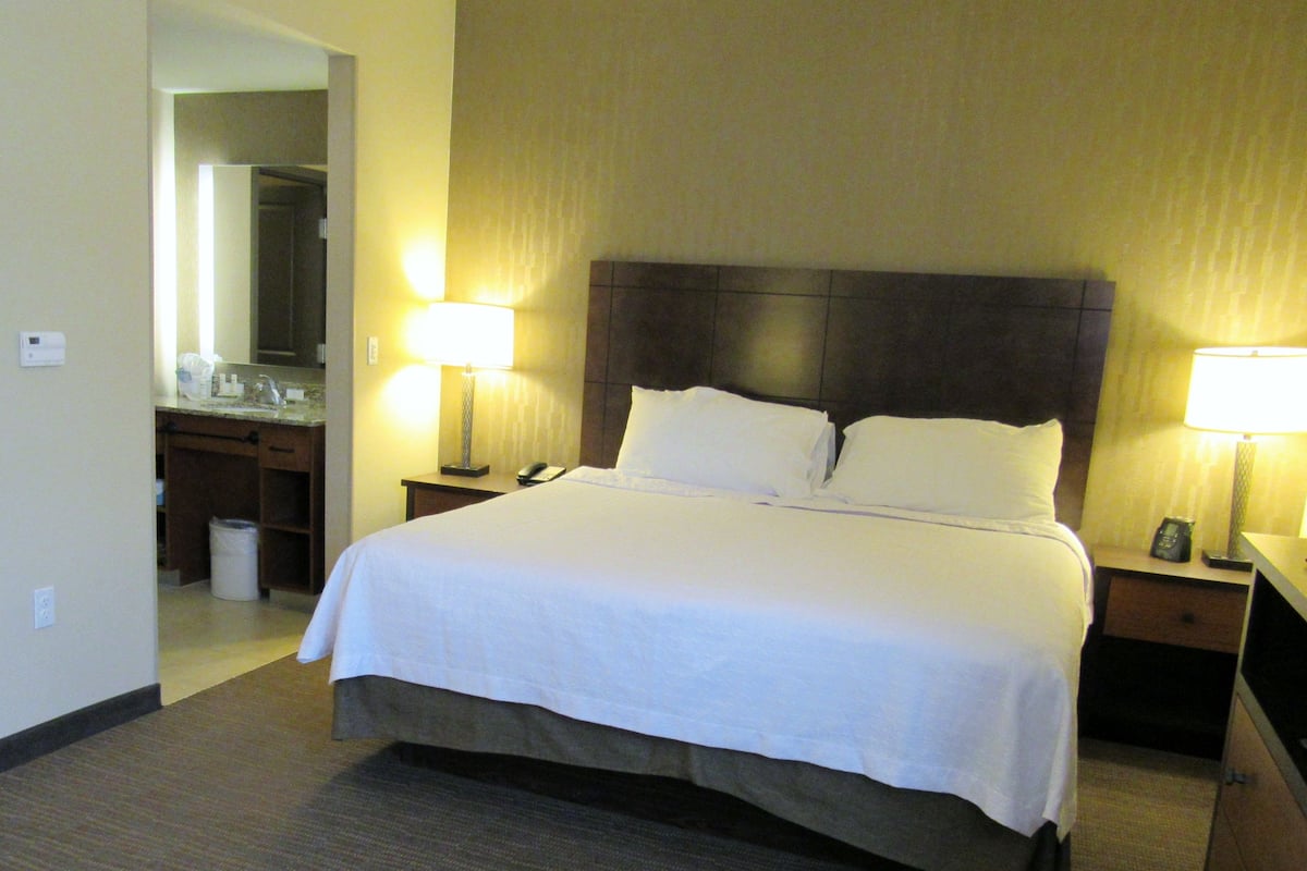 A hotel room with a large bed, two bedside lamps, a nightstand, a mirror, and a bathroom visible through an open door.