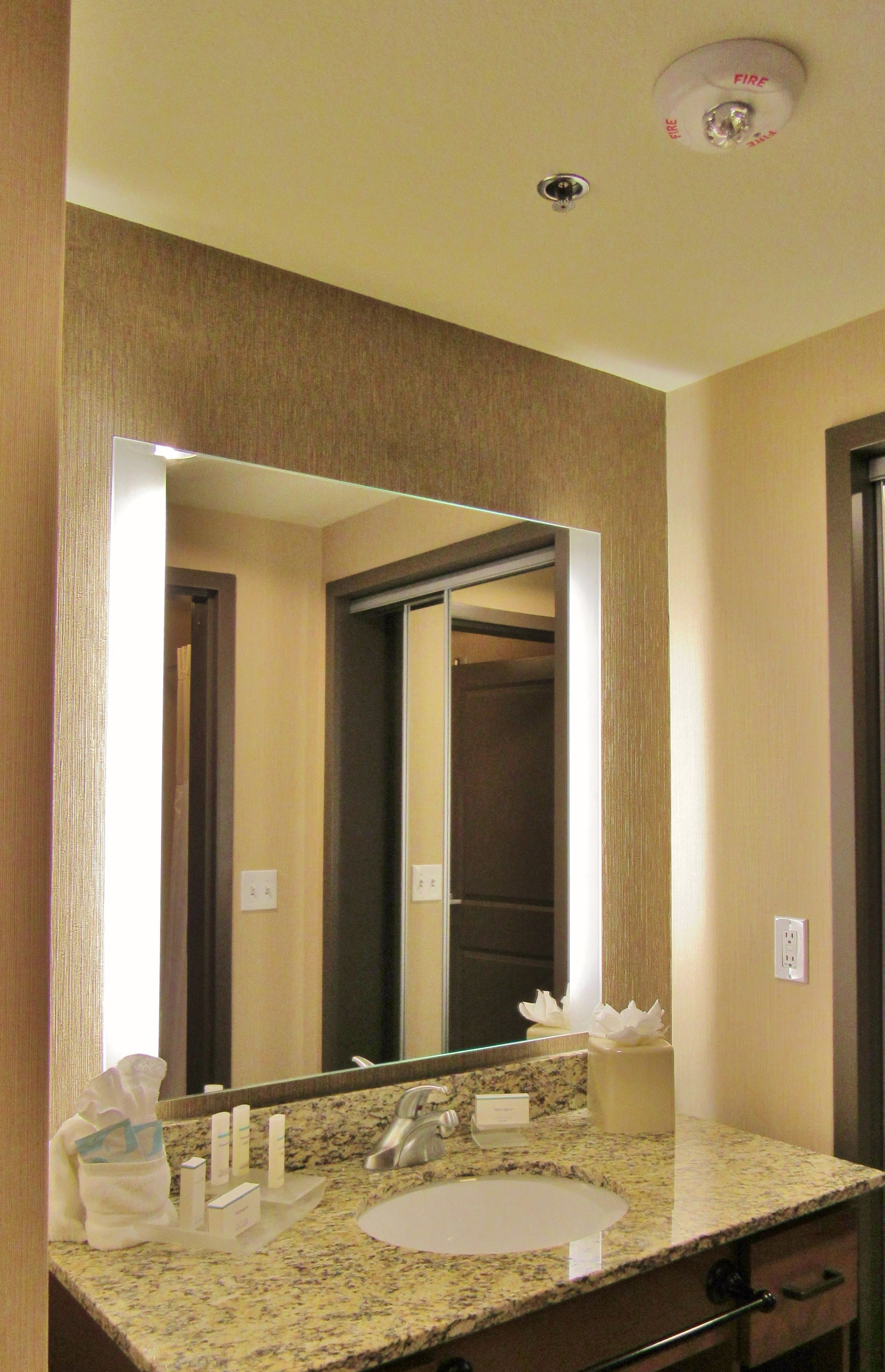 A hotel bathroom with a granite countertop sink, a large mirror, and a visible door leading to another room.