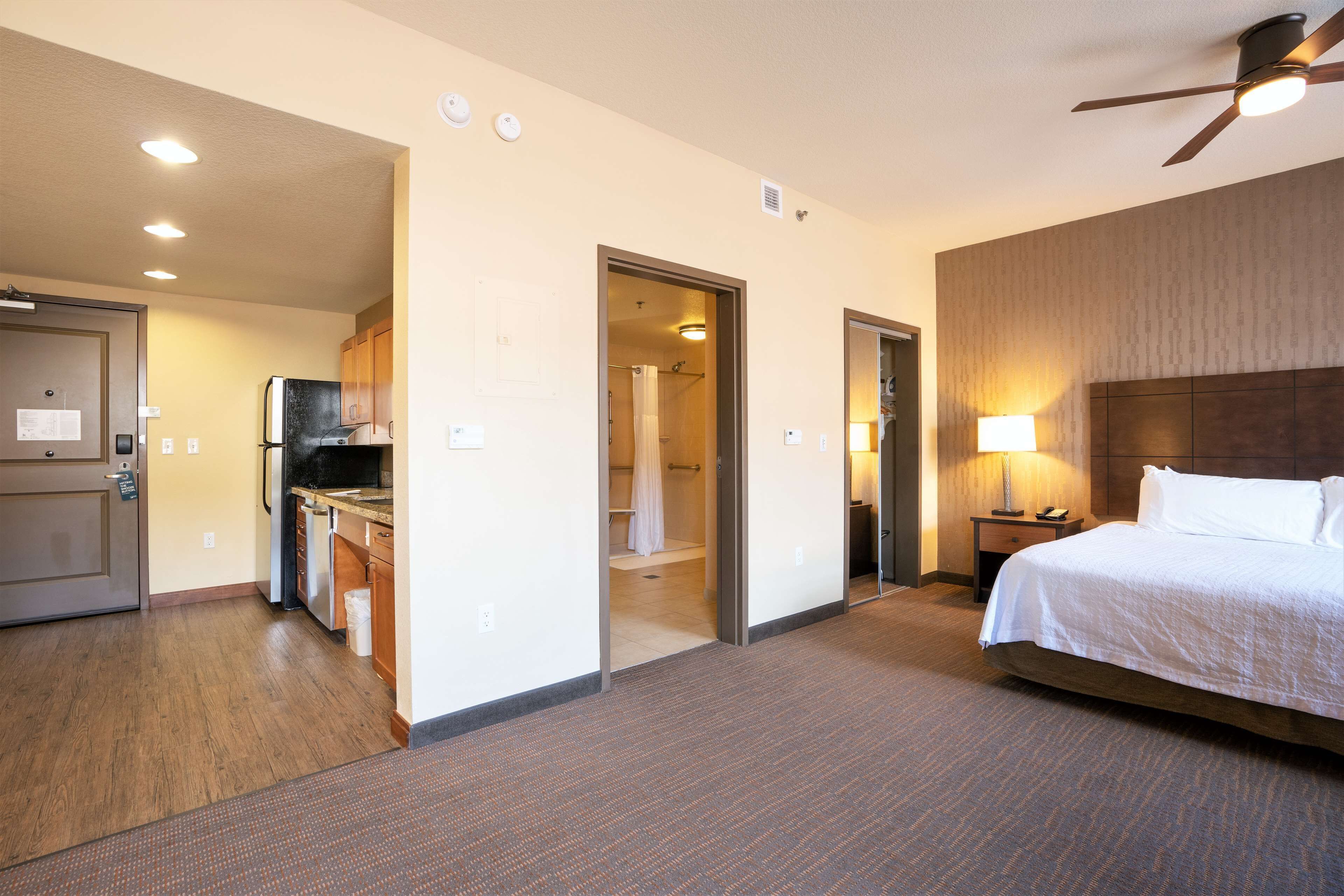 Una habitación de hotel con cama, mesita de noche, lámpara, ventilador de techo, kitchenette y baño.