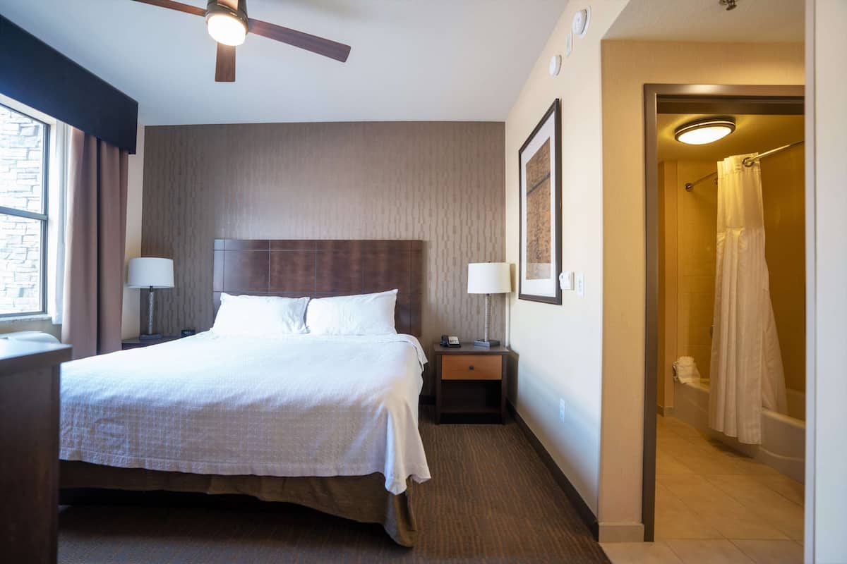 A hotel room with a bed, bedside lamps, a nightstand, a ceiling fan, a framed picture on the wall, and a bathroom with a shower curtain.