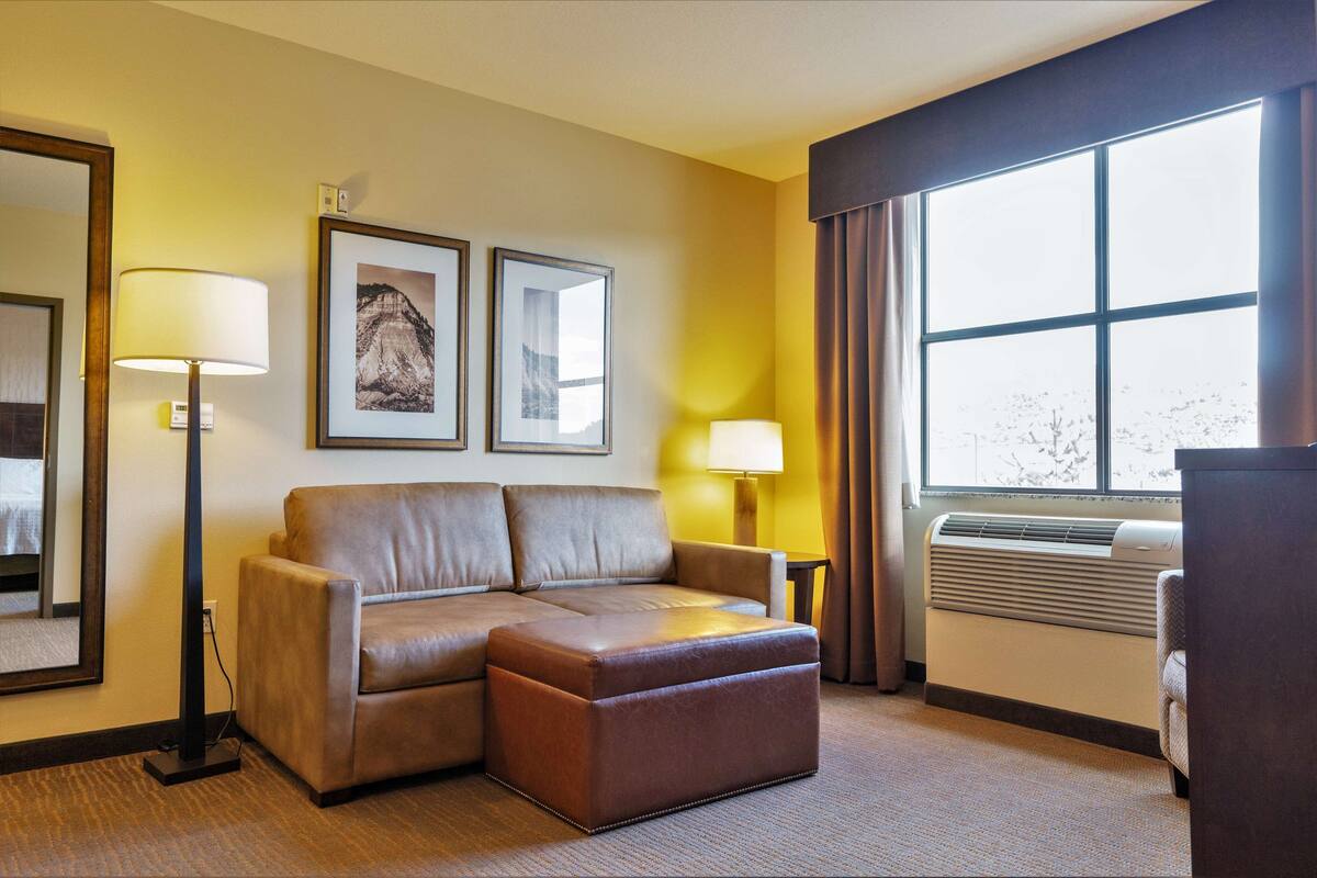 A hotel room with a brown leather sofa, a small ottoman, a standing lamp, a framed picture on the wall, and a large window with curtains.
