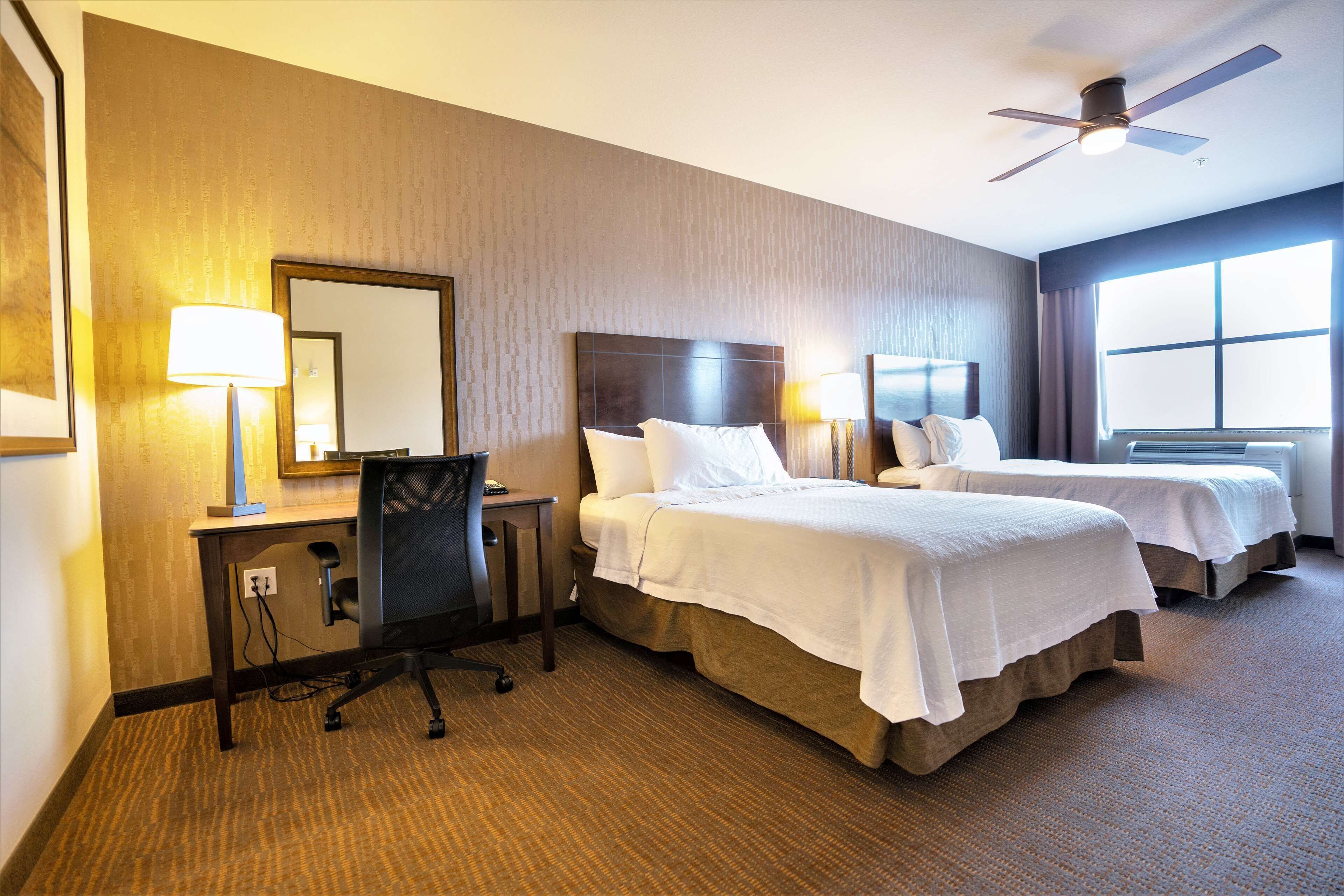 Ein Hotelzimmer mit zwei Betten, einem Schreibtisch mit Stuhl, einem Deckenventilator und einem großen Fenster.