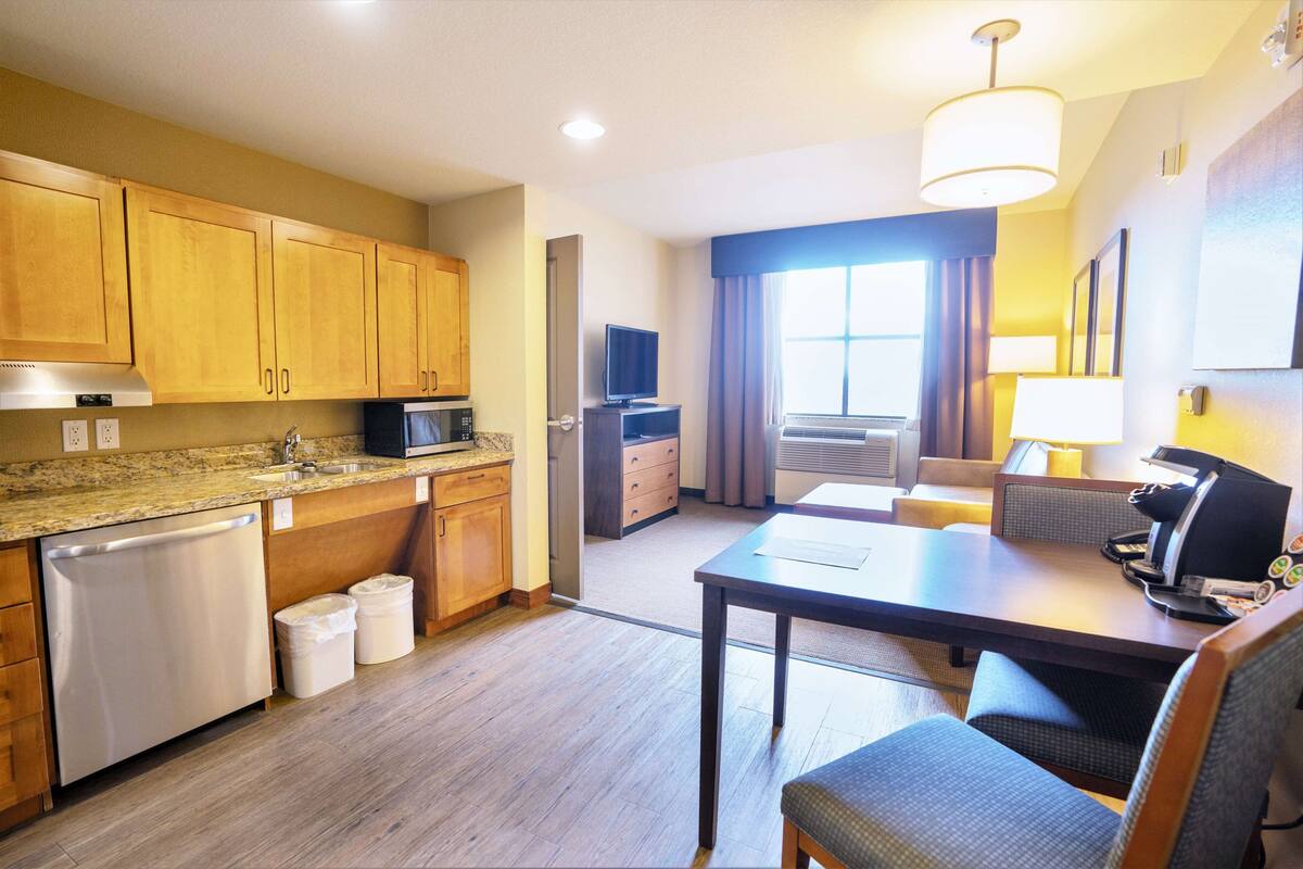 A compact kitchen with wooden cabinets, a microwave, and a dishwasher. There is a dining area with a table and chairs, and a large window with curtains.