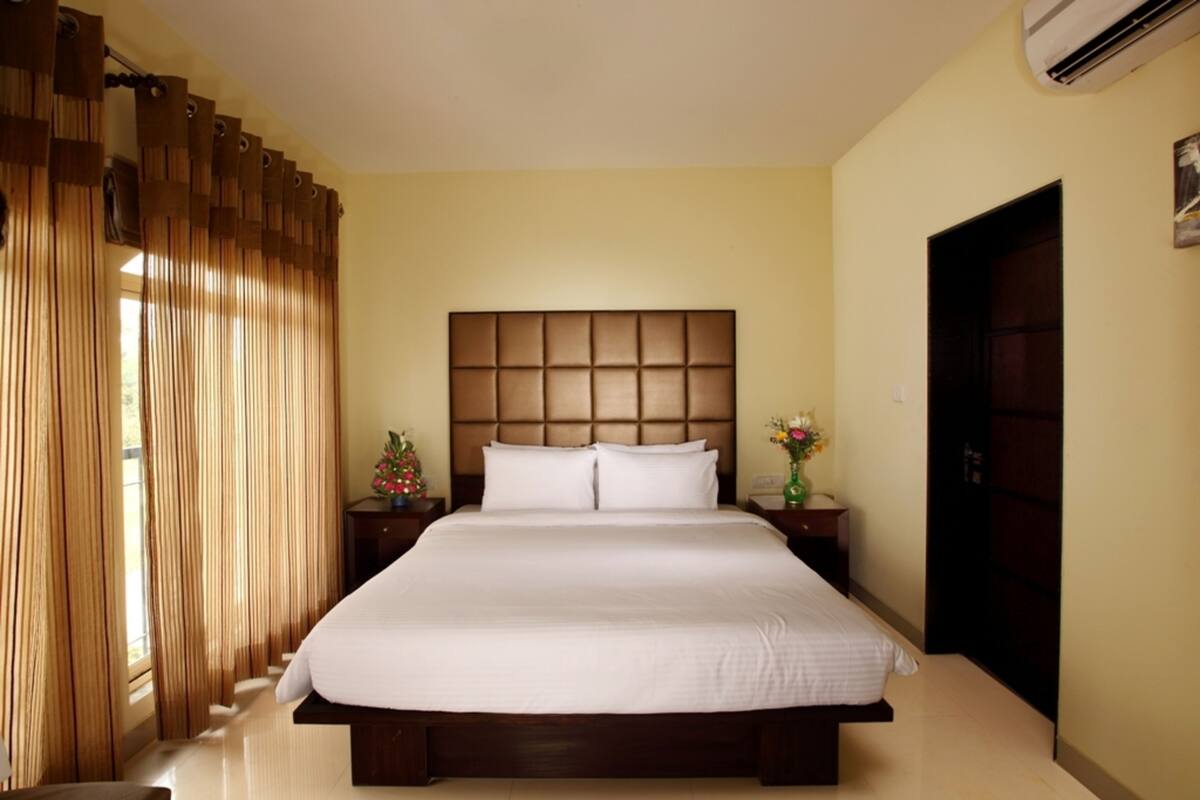 A hotel room with a large bed, wooden headboard, two bedside tables with lamps, a nightstand with a vase of flowers, and a door on the right.