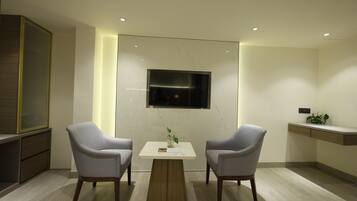 Una habitación moderna con dos sillas grises, una pequeña mesa blanca, un televisor montado en la pared y una planta sobre una mesita auxiliar.