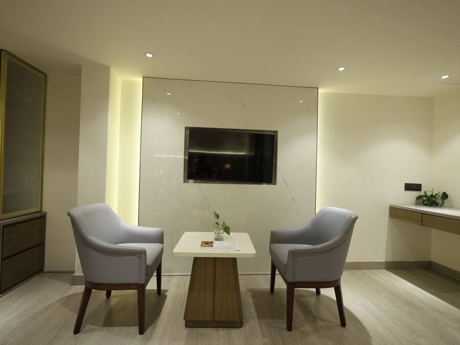 Una habitación moderna con dos sillas grises, una pequeña mesa blanca, un televisor montado en la pared y una planta sobre una mesita auxiliar.
