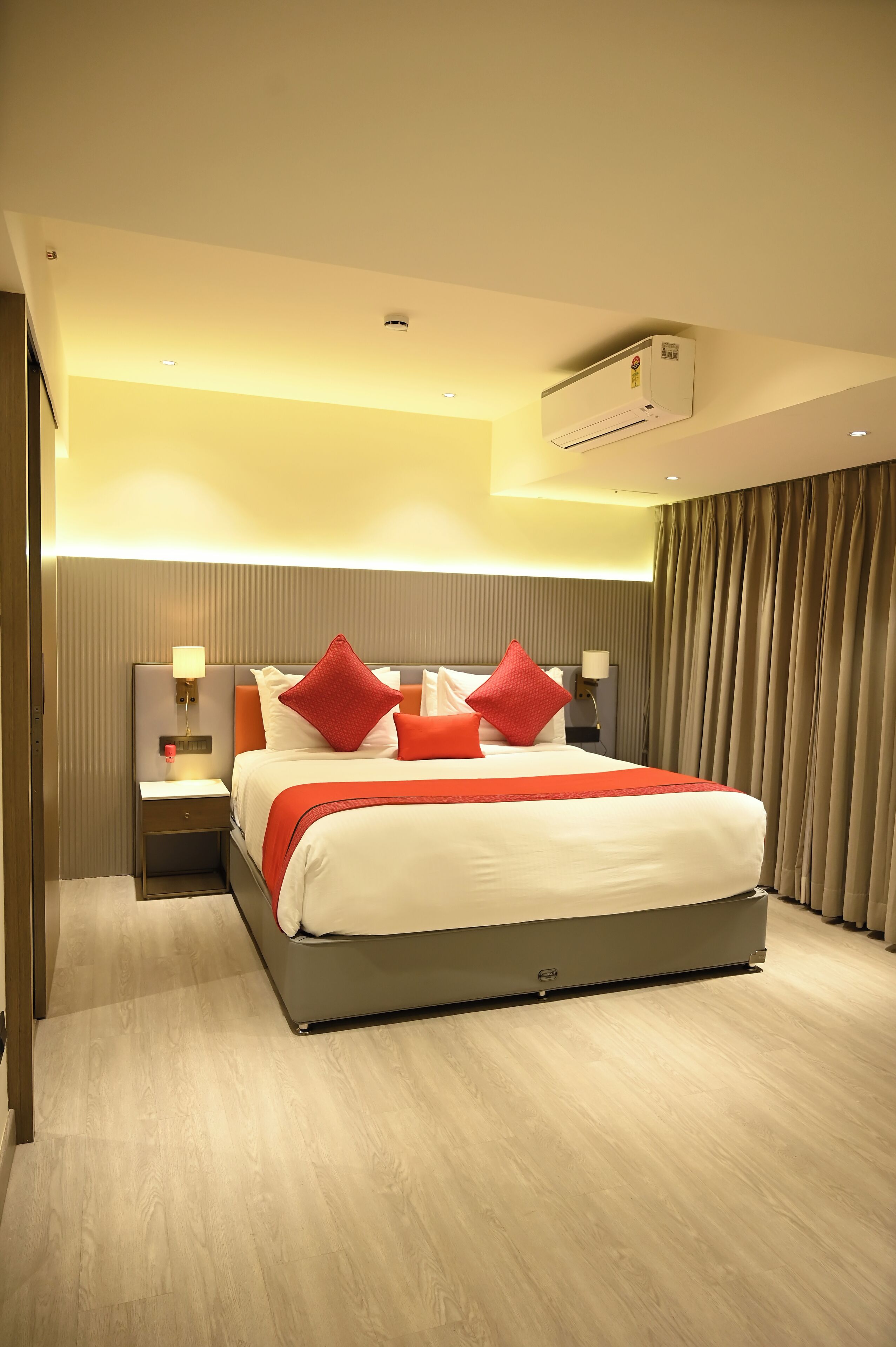 A hotel room with a large bed, a nightstand, a lamp, and an air conditioning unit.