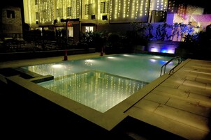 Una piscina con superficie de agua cristalina, rodeada por un solárium empedrado y una pequeña escalera que lleva a un edificio.