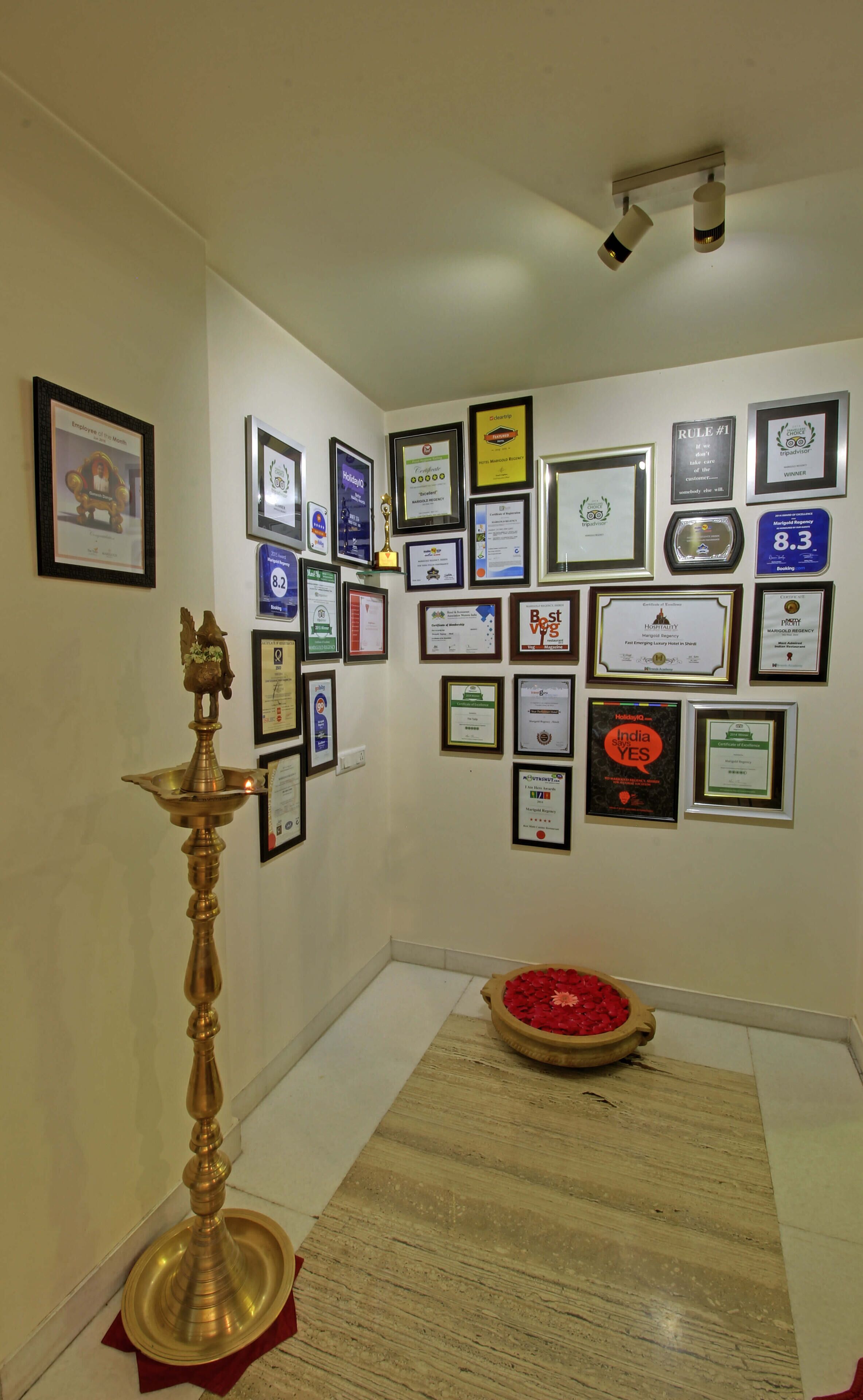 Pared con varios cuadros de premios y certificados, una lámpara de aceite de bronce y un pequeño arreglo floral rojo sobre un piso de madera.