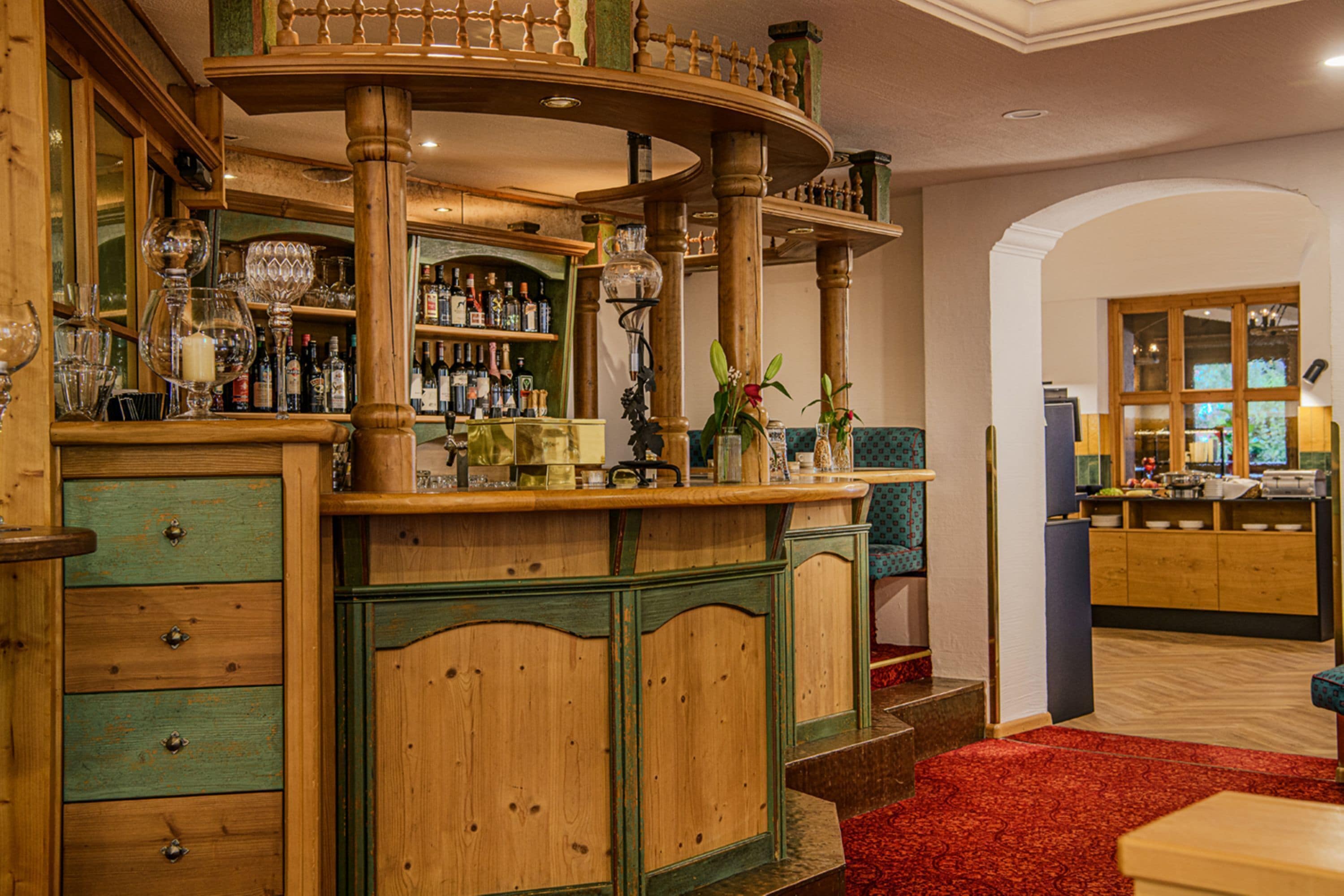 A bar area with wooden furniture, a red carpet, and a kitchen in the background.