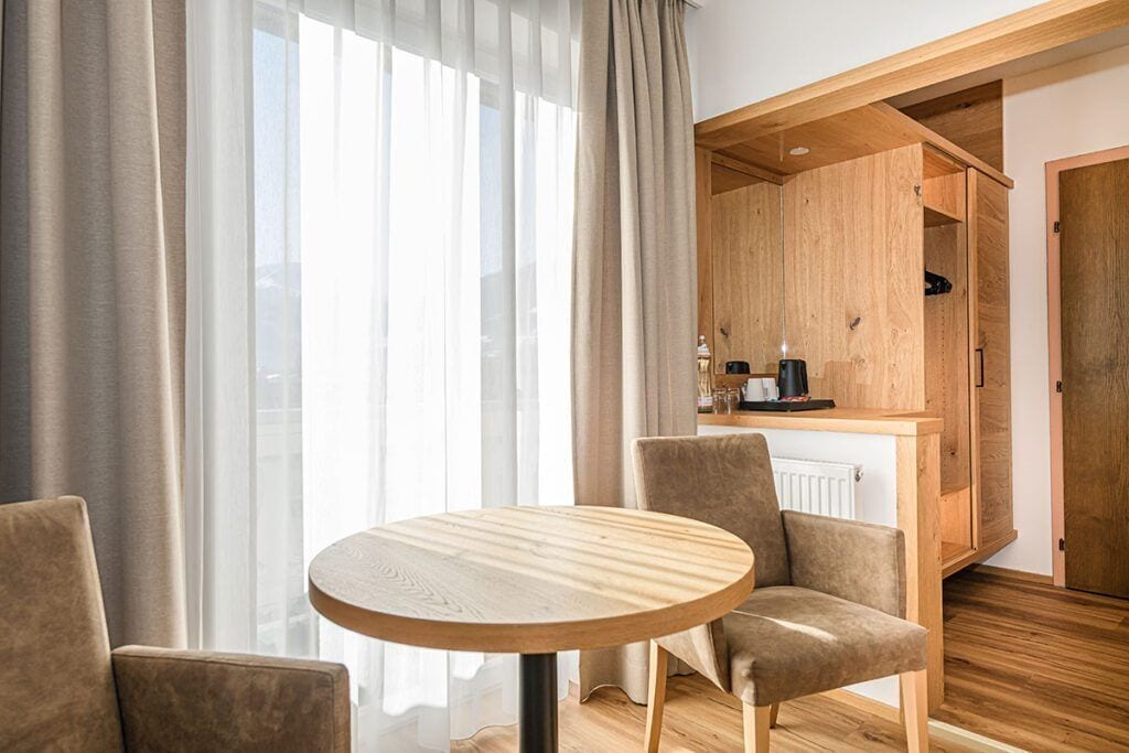 A modern room with a round wooden table, two chairs, a kitchen area with cabinets, and a large window with curtains.